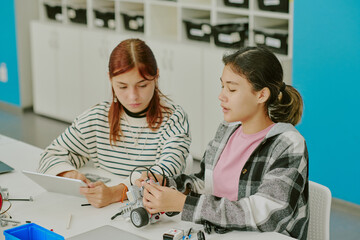 High angle shot of school girl at table making robot on wheels together while red haired girl checking instruction on tablet