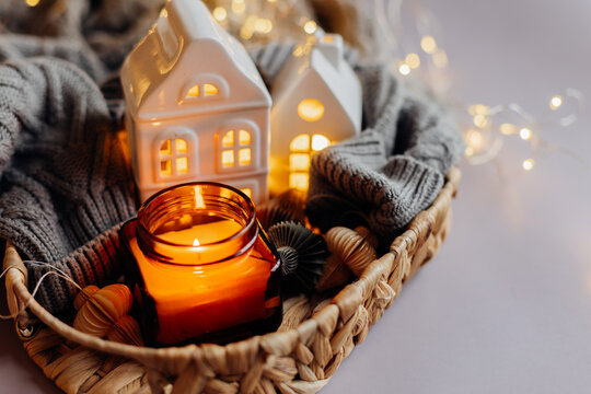 Winter or autumn cozy interior decorations. Burning candle in jar, white decorative ceramic houses, gray cozy sweater and small paper honeycomb baubles. Christmas cozy winter home decor.