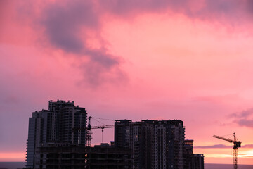 Tall buildings under construction, silhouetted against vivid pink and purple sunset clouds by the sea, highlighting a vibrant urban skyline