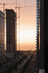 Street bustling with heavy traffic, framed by multi-story buildings and cranes, bathed in sepia tones and backlighting, illustrating the dynamic life of a vibrant metropolitan area