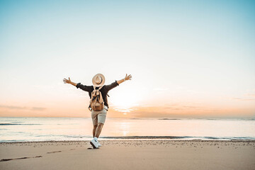 Happy man with backpack standing with arms up at the beach - Delightful tourist enjoying summer vacation by the seaside - Traveling life style and well being concept