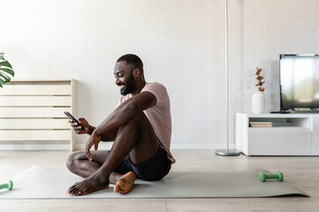 A young man sits on a yoga mat at home after exercising, smiling and using his smartphone, surrounded by plants and natural light in a relaxed atmosphere.