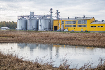 large grain elevator with multiple cylindrical metal silos, structural elements, and a complex system of pipes. It stands against a blue sky, with a field in the foreground after harvest