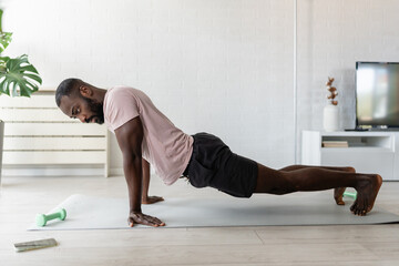 Young African-American man doing a side plank exercise on a yoga mat at home, practicing strength and balance in a bright living room with indoor plants.