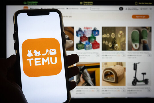 Silhouette of Hand Holding Phone with Temu Logo in Front of Temu Online Shopping Platform Website on Computer Screen