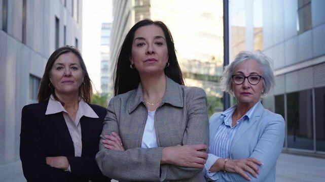 Three mature caucasian empowered businesswomen looking at camera confident. Serious women with cross arms standing outside office district.