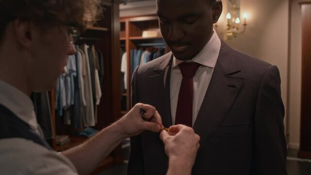 Tilt up shot of sales assistant helping Black male customer to attach tie clip while trying on elegant suit in mens clothing store