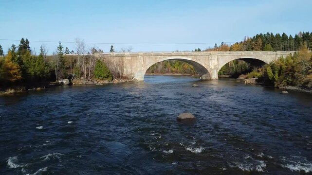 Fast‑moving drone flight over the swirling eddies of the Mitis salmon river, diving toward and passing beneath the concrete arch of the Arthur‑Bergeron road bridge in Grand‑Mitis, , Quebec, Canada
