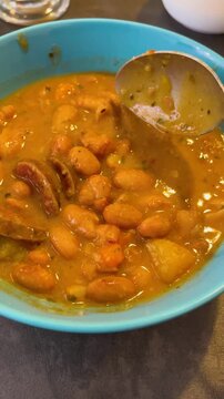 A spoon mixes a thick soup with beans and pieces of meat and sausage in a bowl. A hearty first course for lunch.