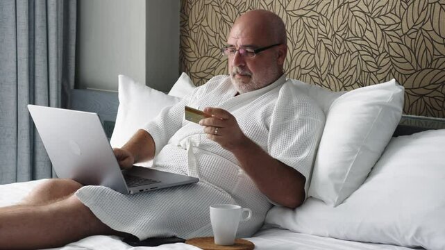 A man in a bathrobe sits on a bed, using his laptop to make online purchases with his credit card. A blend of convenience and relaxation, showcasing the ease of digital living.