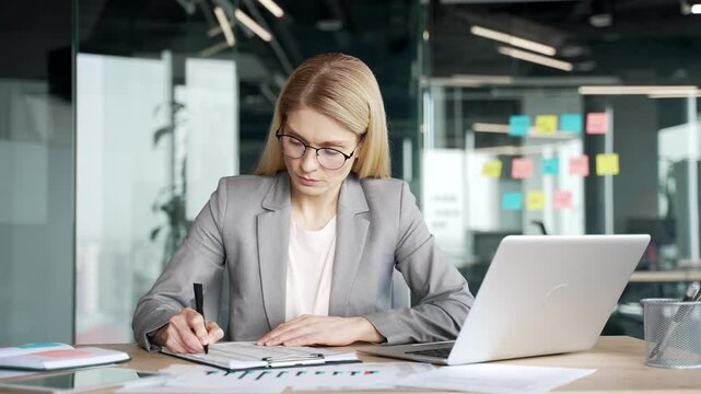 Confident female financier filling out documents with a pen. Accountant is engaged in paper work, making financial calculations using typing on a laptop. Businesswoman in suit fills out blank tax form
