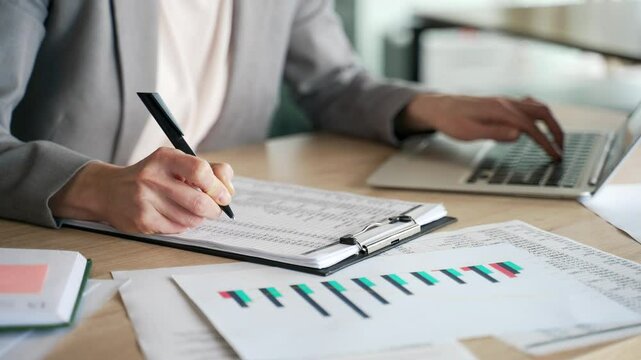 Close up of a female hand filling out documents with a pen. Accountant is engaged in paper work, making financial calculations using typing on laptop. Businesswoman in suit fills out a blank tax form