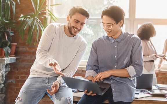 Two young men colleagues sharing creative ideas at workplace, looking at laptop screen, casual office atmosphere