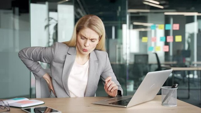 Tired businesswoman in jacket suffering from back pain while working on laptop while sitting at workplace in office. Upset female employee holds her hand on her lower back, massaging her sore muscles