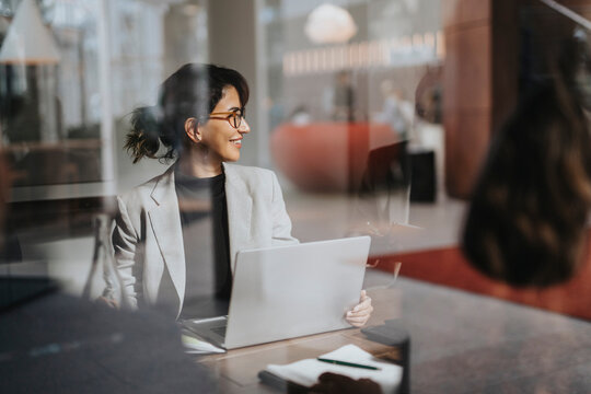 Smiling businesswoman sitting with laptop at desk seen through glass