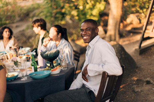 Side view of smiling man sitting on chair with friends at dining table during dinner party