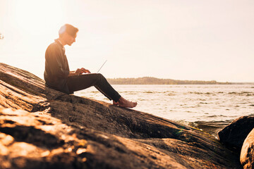 Side view of young man using laptop while sitting on rock near lake at sunset
