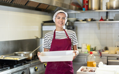 Smiling young female worker wearing striped t-shirt, red apron and cap holding empty tray in...