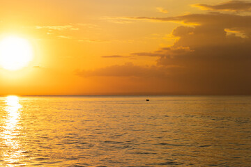 Fototapeta premium This image captures a golden sunset over the ocean, with the sun casting warm light across the water. A small boat is visible on the horizon, adding a peaceful, solitary element to the serene scene.