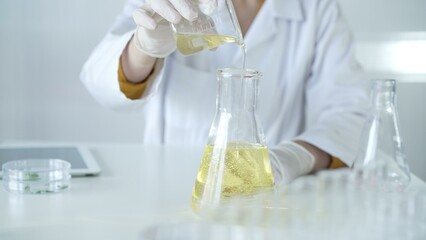 A scientist, wearing a lab coat and white protective gloves, is pouring a yellow oily liquid from one beaker to another in laboratory, close up. Medicine and science concept