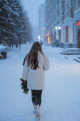 A woman dressed in a white coat is walking leisurely down a snowy street, where everything is covered in a blanket of white snowflakes