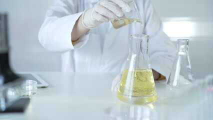 A scientist, wearing a lab coat and white protective gloves, is pouring a yellow oily liquid from one beaker to another in laboratory, close up. Medicine and science concept