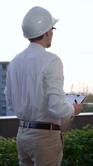 Man a constructive engineer wearing white shirt and hard hat is making notes on a clipboard while inspecting a building site at sunrise, back and vertical view . Architect at work