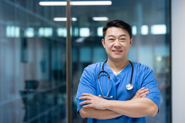 Confident asian male doctor wearing blue scrubs and stethoscope, standing in modern hospital environment with arms crossed, smiles warmly indicating professionalism and care.