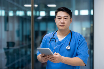 Asian doctor in blue scrubs with stethoscope using digital tablet in modern hospital. Focused...