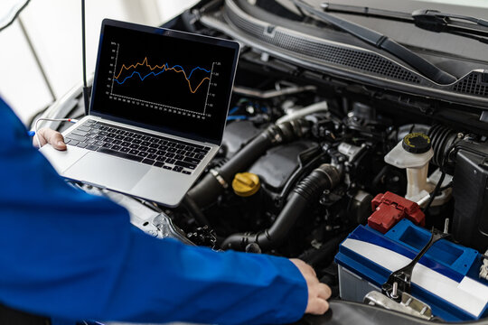 An automotive technician analyzes data using a laptop while inspecting an engine in a workshop during the day