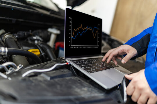 Mechanic using a laptop to diagnose issues under the hood of a vehicle in a garage during daylight hours