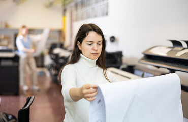 Woman, printing establishment worker, examining large piece of paper, checking results of printing.