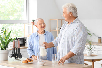 Happy senior couple with tasty buns pouring milk from jug into glass in kitchen