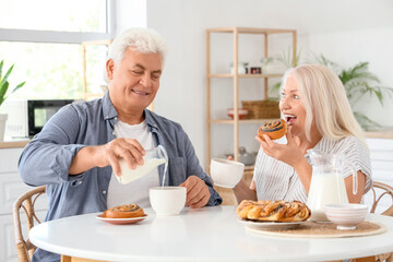 Happy senior couple with tasty buns pouring milk into cups in kitchen
