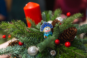 A person is delicately holding a beautifully arranged Christmas decoration that features a vibrant red candle, pine cones, berries, and festive decorations