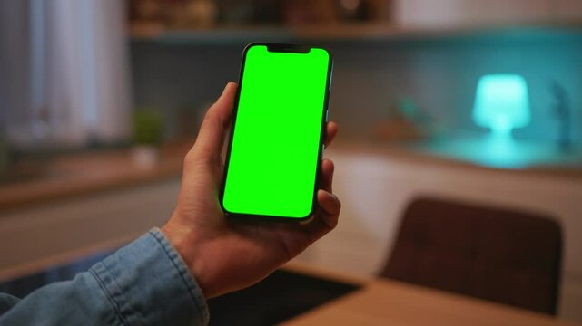 Man's left hand holiding smartphone with green screen in vertical position isolated on blurred background of modern kitchen. Mock up mobile screen. Modern technologies. Indoors. Man using gadget