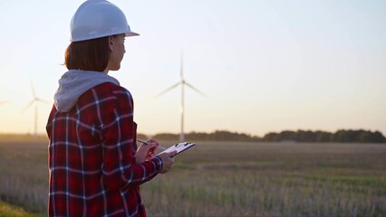 Adult woman engineer wearing white cask is taking notes on a clipboard on a field with wind turbines