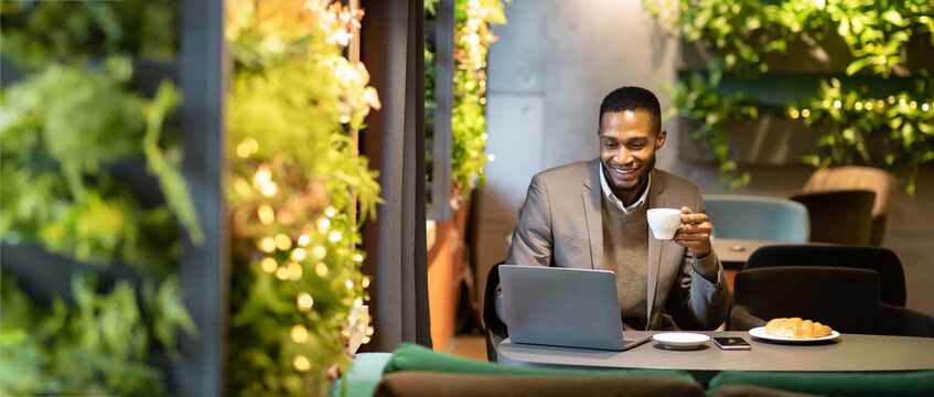 Adult black man holding white mug, smiling and looking at open laptop computer on table at cafe, copyspace