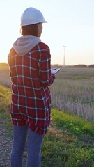 Woman engineer wearing a white protective helmet is taking notes with a clipboard in a field with wind turbines, as the sun sets. Clean energy and engineering concept