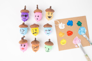 Painted acorns with faces, various colors, 3x3 grid, white background, brown caps, happy, sad, surprised expressions, creative autumn craft, whimsical nature art, decoration,