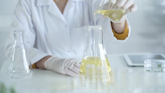 A scientist, wearing a lab coat and white protective gloves, is pouring a yellow oily liquid from one beaker to another in laboratory, close up. Medicine and science concept