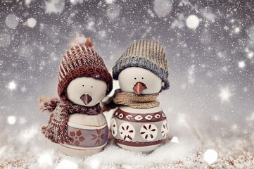 Two adorable snowbird figurines dressed in cozy knit hats and scarves, standing side by side in a snowy setting with sparkling bokeh lights, creating a warm and festive holiday scene.