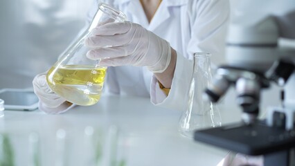 Microscope is near a scientist, wearing a lab coat and white protective gloves, and holding beaker...