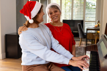 Senior female friends wearing Santa hats playing piano and laughing at home