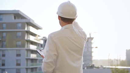 Man construction engineer wearing white shirt and hard hat is making notes on a clipboard while inspecting a building site at sunset, back view. Architecture and engineering