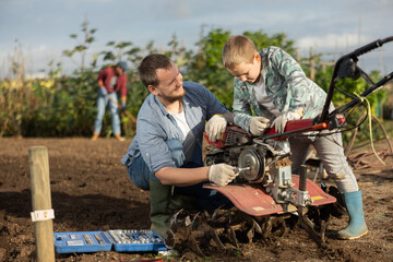 Male farmer shows his son how to fix garden cultivator. Farmer and a guy are repairing a machine to...