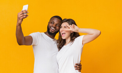 Cheerful interracial couple taking selfie with smartphone, having fun together over yellow background. Playful girl showing peace gesture, empty space