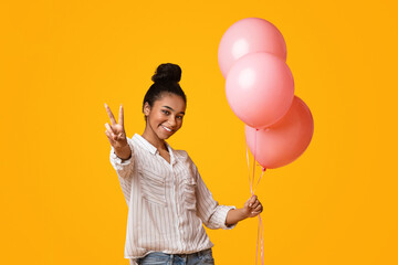 Portrait of laughing afro girl with pink air balloons showing peace sign while posing on yellow...