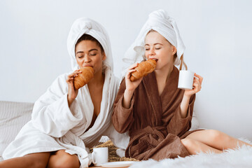 Joyful multiethnic spa girlfriends having breakfast together, eating fresh croissants, drinking...