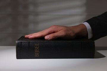 Man taking oath with his hand on Bible at white table, closeup